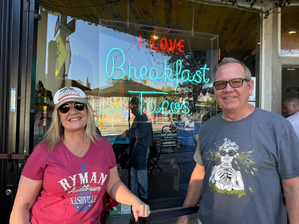 Two smiling guests in front of a neon sign that reads "I LOVE Breakfast Tacos" on To Live and Dine in LA Food Tours' Los Angeles Taco Tour.