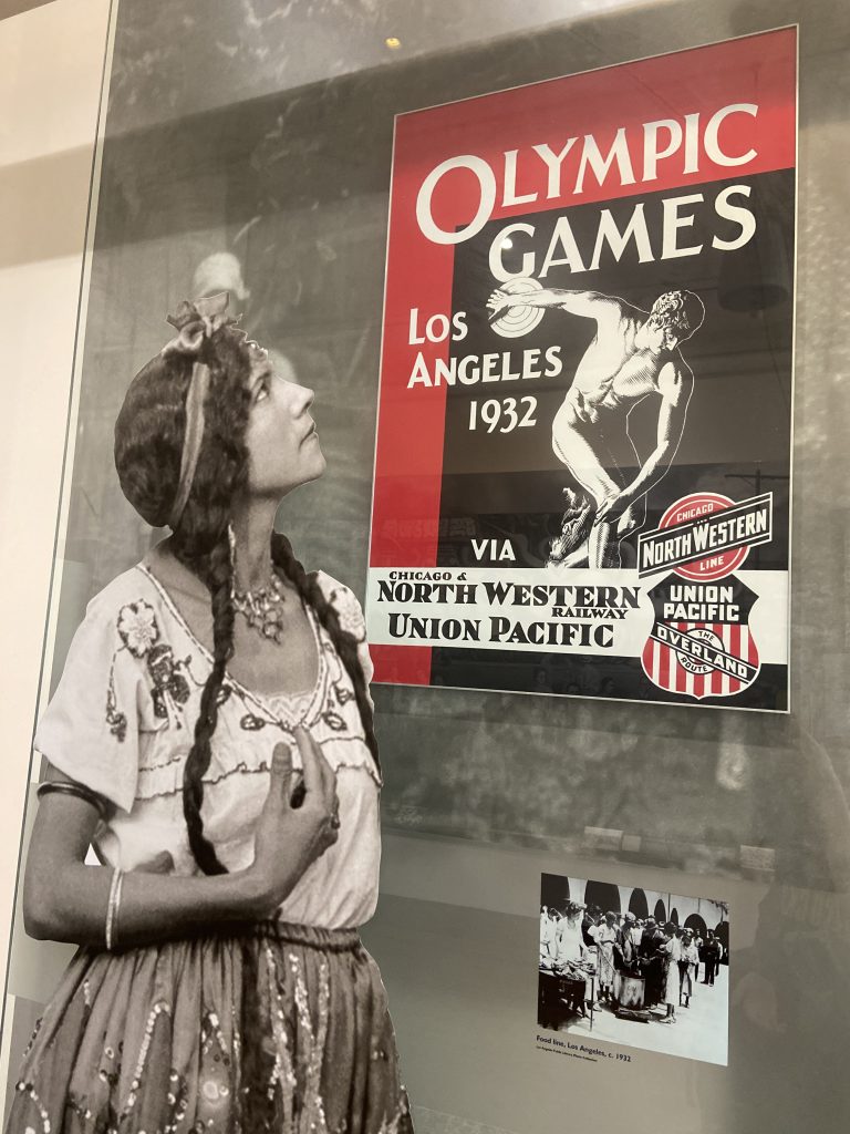 A woman from the 1920s in traditional Mexican dress looks at an Olympic Games Los Angeles 1932 poster printed by Chicago & Northwestern and Union Pacific Railways in downtown Los Angeles on To Live and Dine in LA Food Tours' Chinatown and Olvera Street Food Tour.