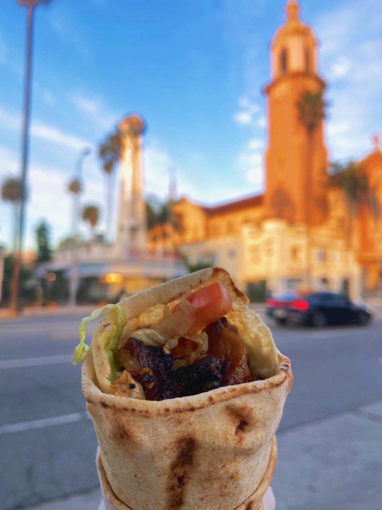A chicken shawarma sandwich with toasted pita bread, fresh tomato, and crisp lettuce in the foreground on a Hollywood Food Tour led by To Live and Dine in LA Food Tours. In the background, the
Church of the Blessed Sacrament and Crossroads of the World on Sunset Boulevard in Hollywood, Los Angeles. 